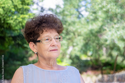 An elderly woman in glasses sits in nature and looks at the camera. The concept of old age, wisdom and joy.