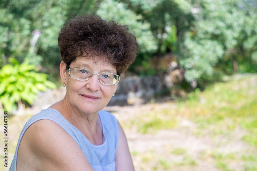 An elderly woman in glasses sits in nature and looks at the camera. The concept of old age, wisdom and joy.