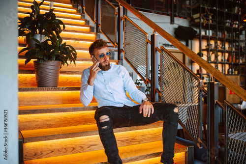 A young caucasian man with sunglasses sitting on the steps in a cafe is talking on the phone. Handsome businessman in a restaurant © Dragica