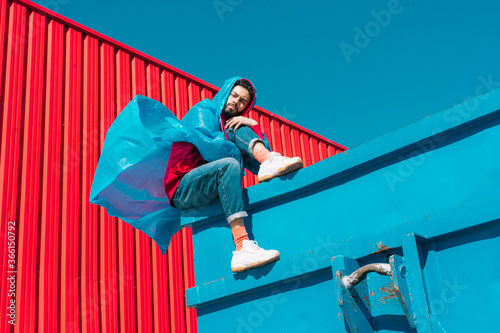 Young man wearing rain coat sitting on edge of blue container in front of red wall photo