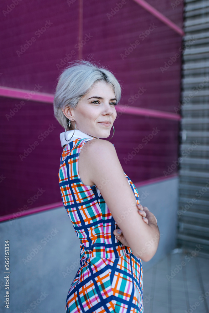 Thoughtful young woman with arms crossed looking away while standing against pink glass wall Thoughtful young woman with arms crossed looking away while standing against pink glass wall