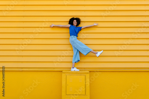Happy young woman balancing on platform in front of yellow wall