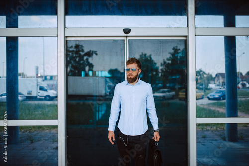 A young caucasian businessman with sunglasses is standing in front of the airport with a black briefcase in his hand