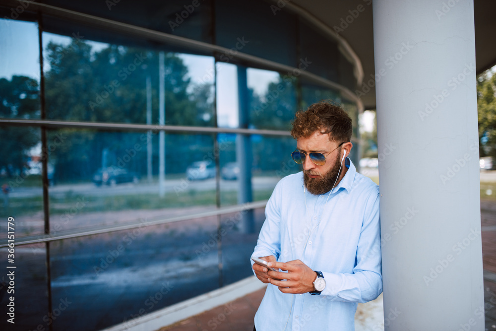 Fototapeta premium A young handsome caucasian businessman with sunglasses stands in front of the company and types a message on the phone