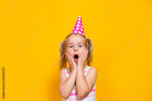 Happy birthday child girl in pink cap on colored yellow background. Toddler holding her mouth with hands.