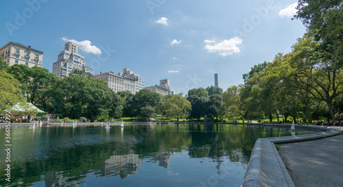 Fototapeta Naklejka Na Ścianę i Meble -  Panoramic image of central park model boat sailing during summer with mirror effect on water reflecting sky, trees and building.
