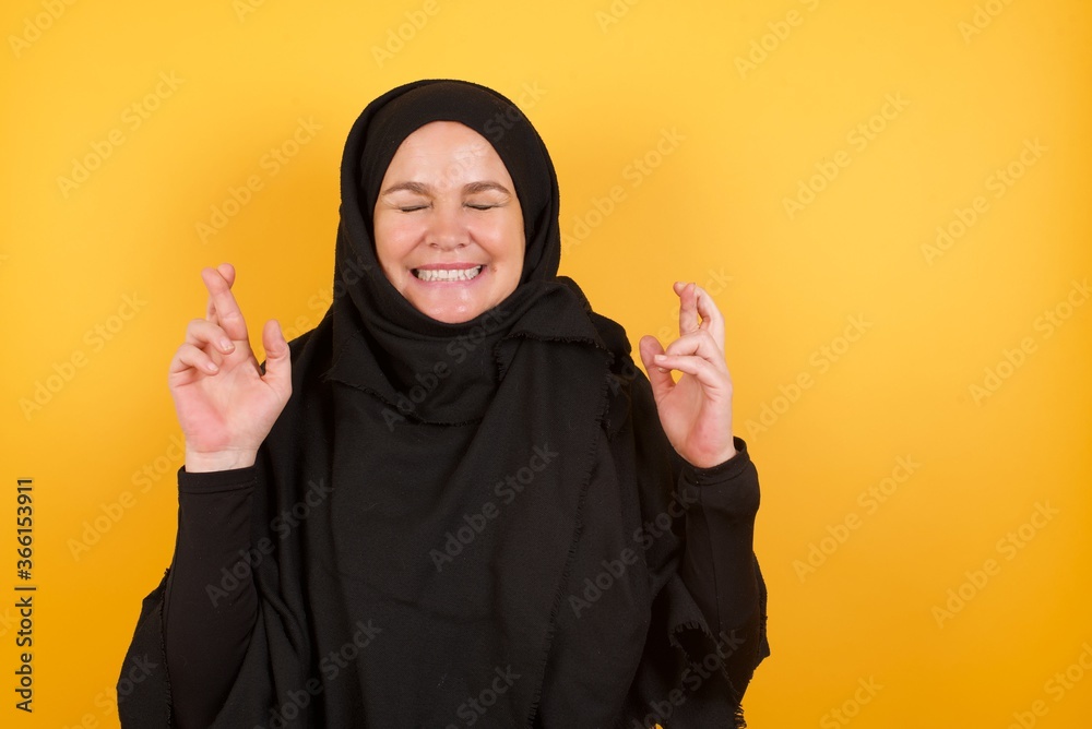 Beautiful middle aged muslim woman wearing black hijab over yellow background. has big hope, crosses fingers, believes in good fortune, smiles broadly. People and wish concept Beautiful middle aged muslim woman wearing black hijab over yellow background. has big hope, crosses fingers, believes in good fortune, smiles broadly. People and wish concept