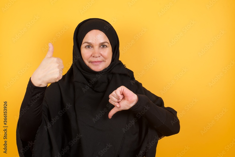 Beautiful middle aged muslim woman wearing black hijab over yellow background showing thumbs up and thumbs down, difficult choose concept