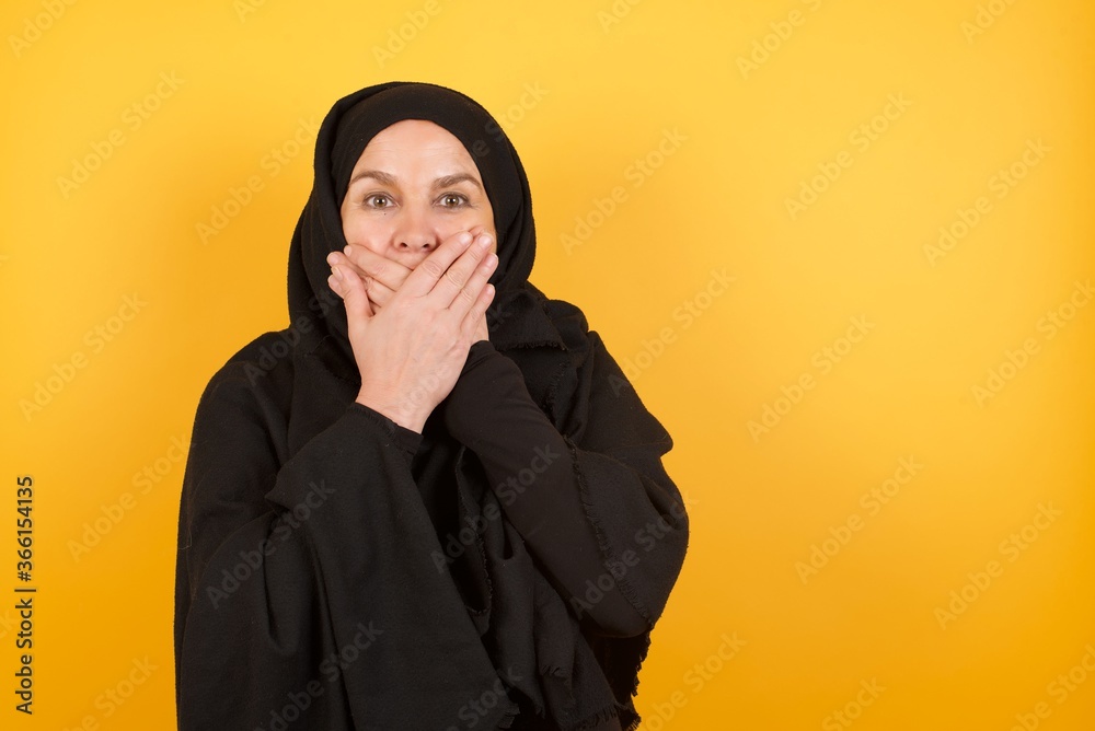 Beautiful middle aged muslim woman wearing black hijab over yellow background shocked covering mouth with hands for mistake. Secret concept.