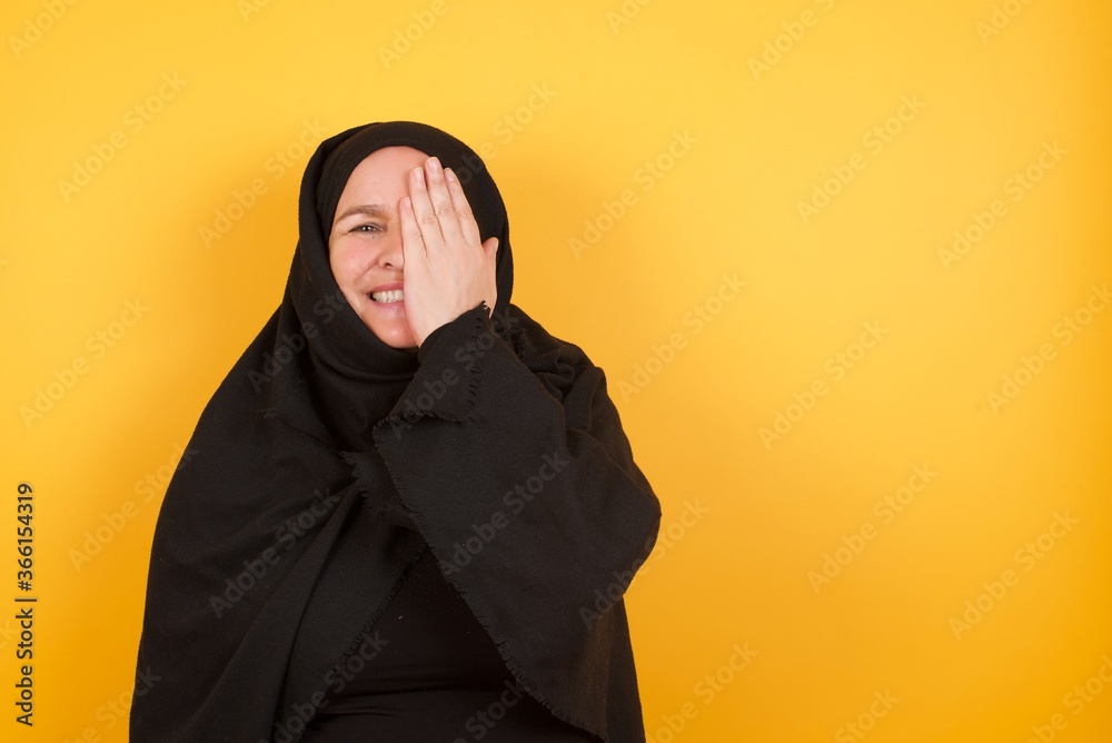 Beautiful middle aged muslim woman wearing black hijab over yellow background covering one eye with her hand, confident smile on face and surprise emotion. Beautiful middle aged muslim woman wearing black hijab over yellow background covering one eye with her hand, confident smile on face and surprise emotion.