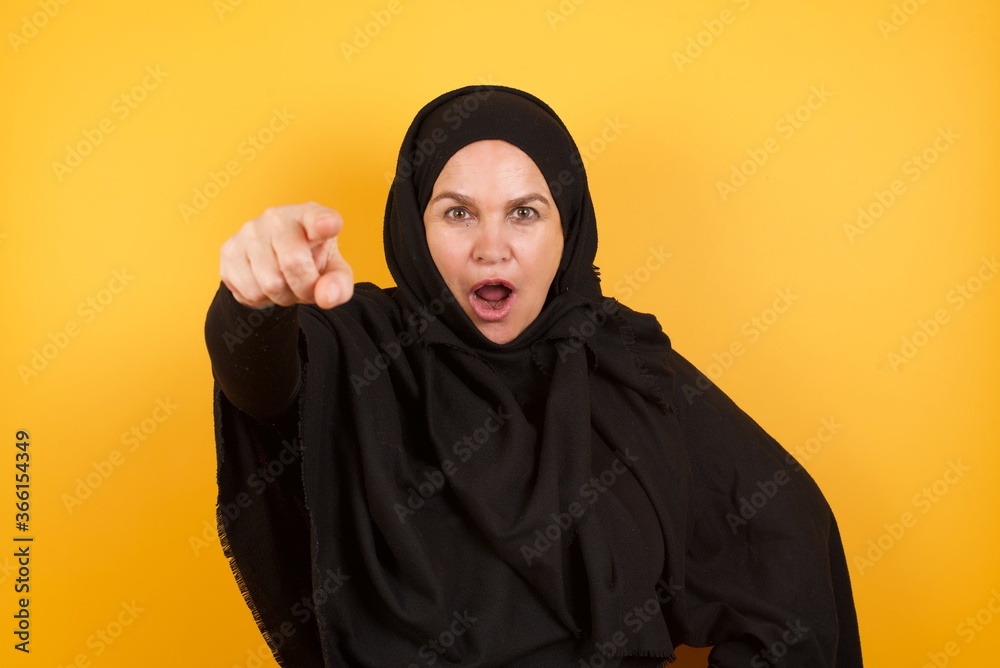 Beautiful middle aged muslim woman wearing black hijab over yellow background Pointing with finger surprised ahead, open mouth amazed expression, something on the front. Beautiful middle aged muslim woman wearing black hijab over yellow background Pointing with finger surprised ahead, open mouth amazed expression, something on the front.