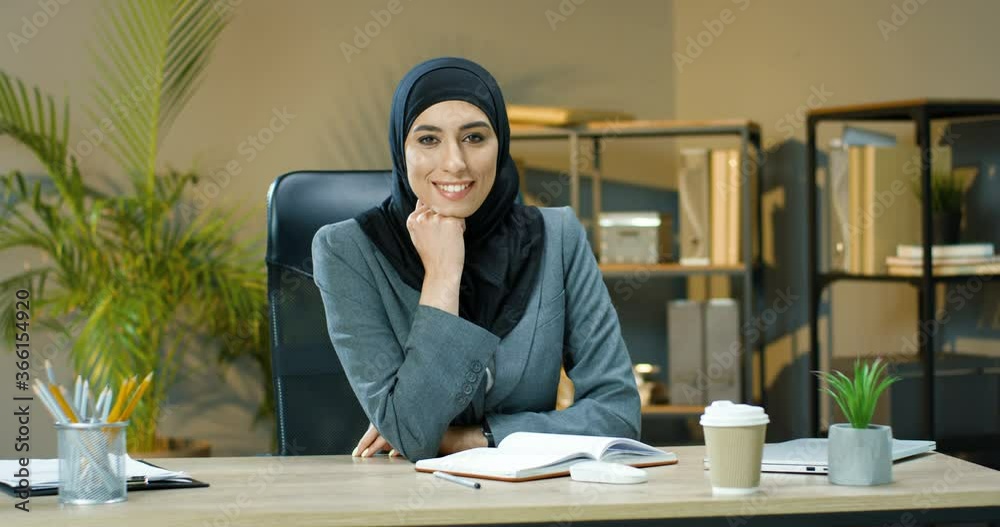 Portrait of pretty happy muslim woman in hijab sitting in office with open notebook on table and looking at camera. Arabian businesswoman in headscarf with smile at desk with planner Business planning Portrait of pretty happy muslim woman in hijab sitting in office with open notebook on table and looking at camera. Arabian businesswoman in headscarf with smile at desk with planner Business planning
