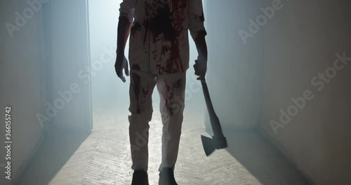 Bloodstained maniac walking in hallway of old house. Bloodthirsty butcher wearing white stained apron and carrying an axe, escaping after murder 4k footage photo