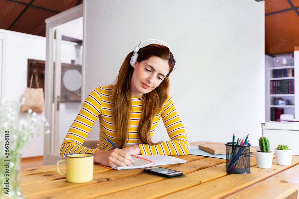 Woman wearing headphones writing in book while looking at smart phone on desk Woman wearing headphones writing in book while looking at smart phone on desk