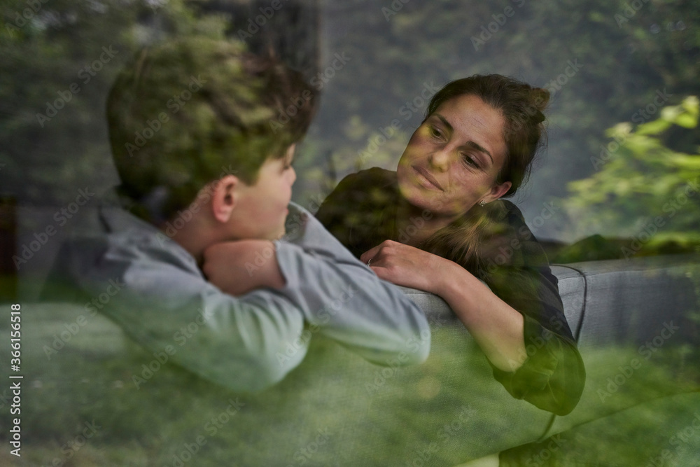 Mother and son talking while relaxing on sofa at home seen through window Mother and son talking while relaxing on sofa at home seen through window