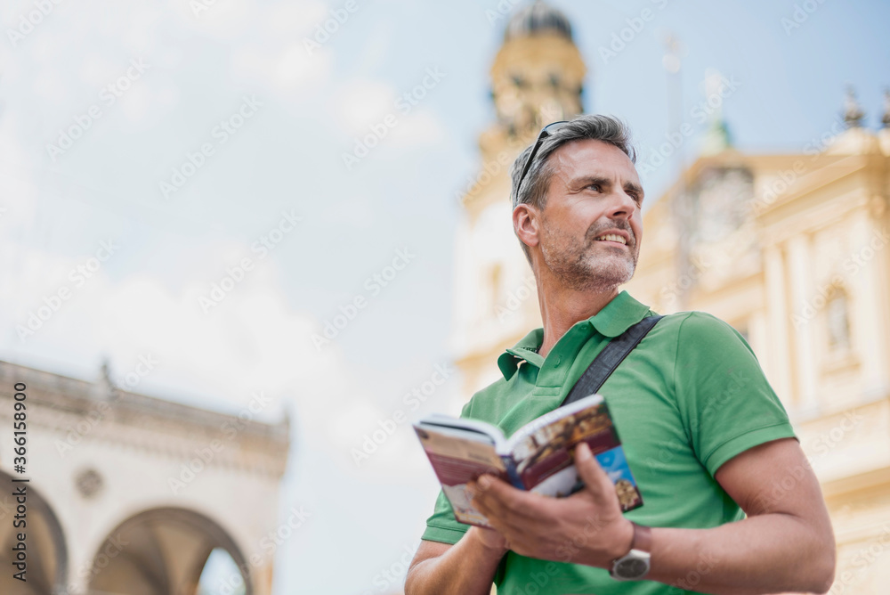 © Westend61 - Smiling mature man holding guidebook looking away while standing in city