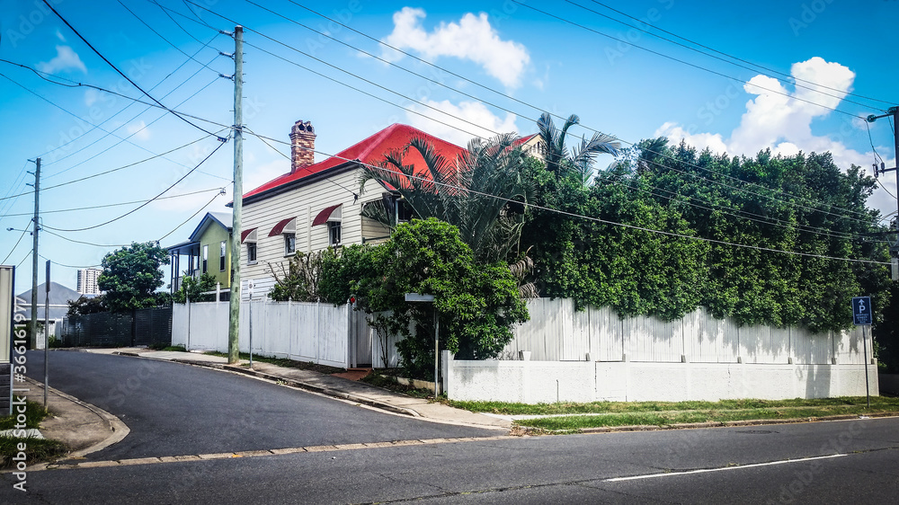 Street corner in Brisbane suburbs with red-roofed house surrounded by white picket fence an shrubbery and  tall building in distance