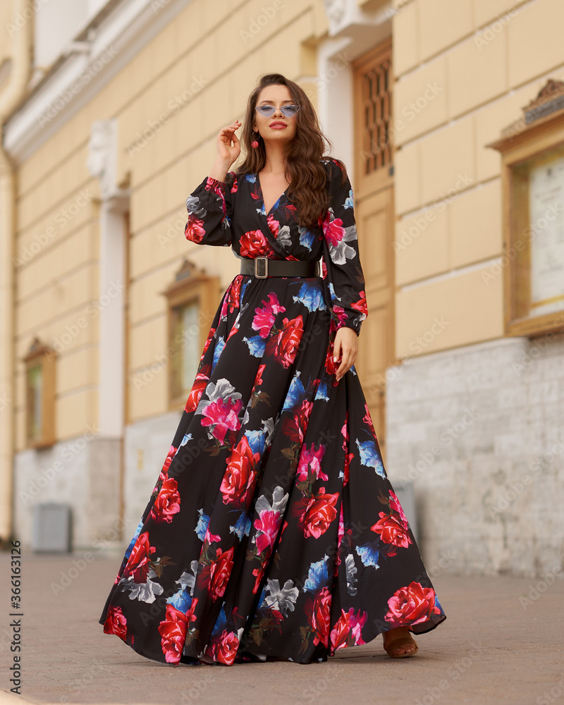 Young beautiful lady wearing black dress with colorful floral design and standing at city street. Pretty woman outdoor full length portrait. Fashion model.