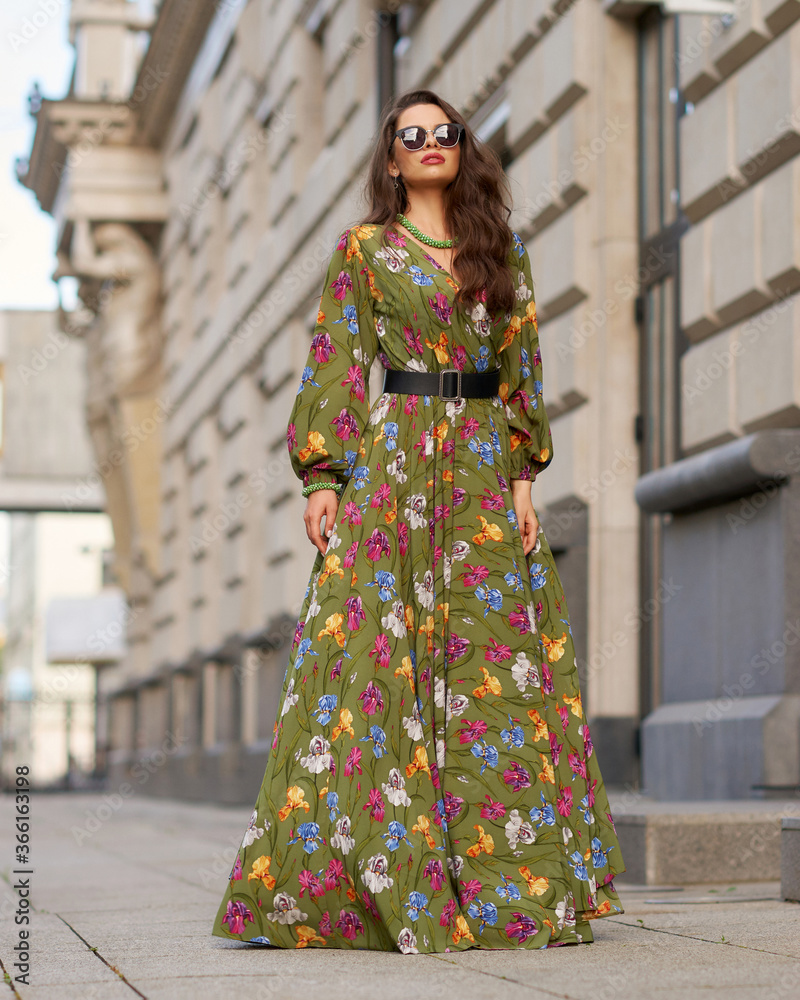 Full length outdoor portrait of young elegant woman wearing green dress with floral design and walking at city street on a summer day