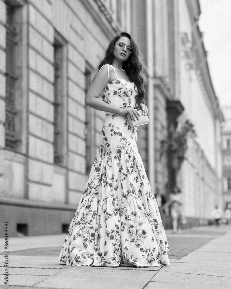 Young beautiful woman with long brunette wavy hair walking at city street. Elegant female wearing long colorful white and red floral dress. Girl in ball gown. Full length fashion portrait Young beautiful woman with long brunette wavy hair walking at city street. Elegant female wearing long colorful white and red floral dress. Girl in ball gown. Full length fashion portrait