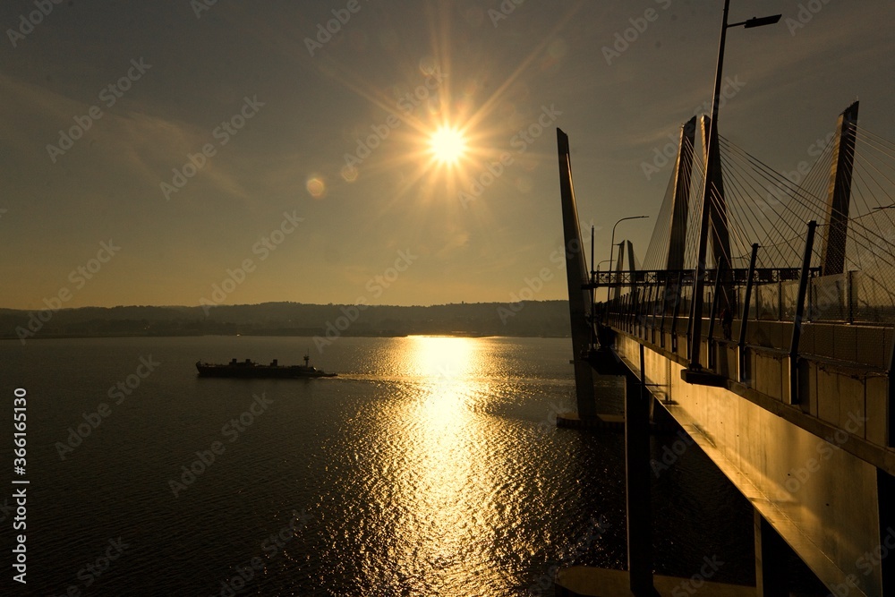 Obraz premium Tarrytown, NY / USA: A landscape image of sunrise at the Governor Mario M. Cuomo Bridge. A silhouette of a ship passing by the bridge, with Tarrytown in the background.