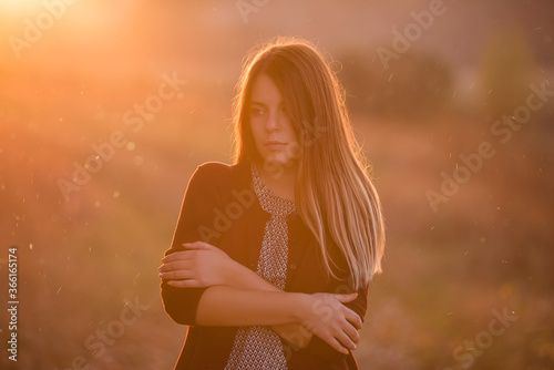 A modern, beautiful girl, in a burgundy jacket, dark jeans, stands in a field, blonde hair fluttering in the wind against the background of an autumn field at sunset. Autumn travel, rest from the city