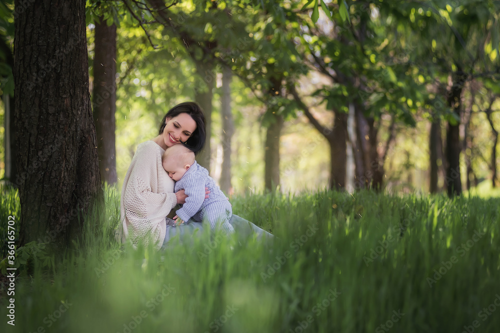 Modern beautiful young brunette mother with a short haircut, holding a toddler in a gray knitted romper in her arms. A girl and her son are sitting under a tree, playing together, hugging. Mom kisses Modern beautiful young brunette mother with a short haircut, holding a toddler in a gray knitted romper in her arms. A girl and her son are sitting under a tree, playing together, hugging. Mom kisses