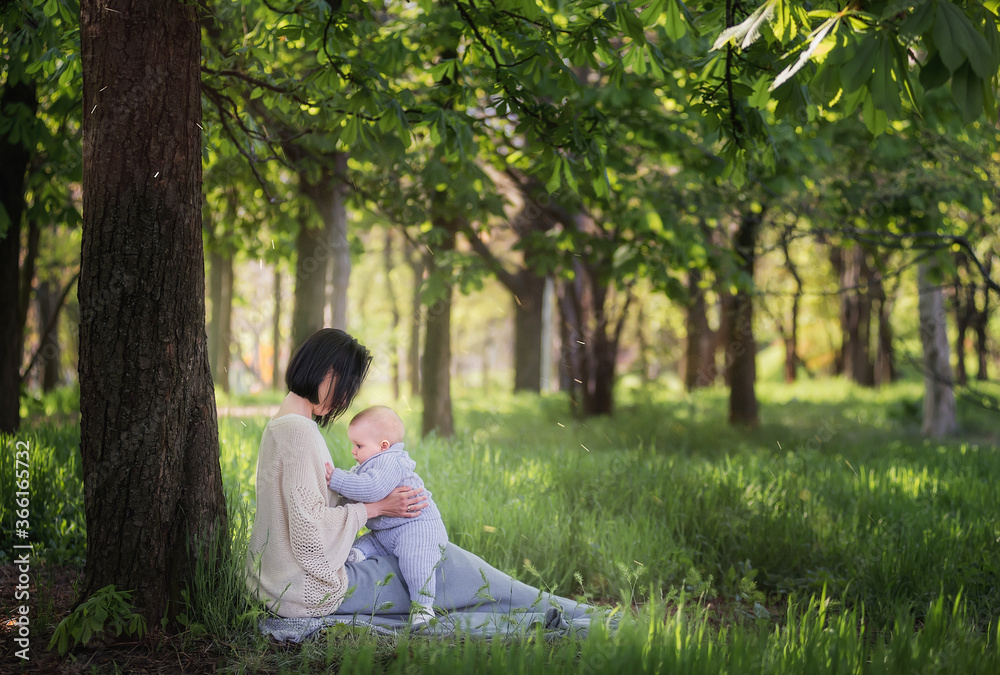 Modern beautiful young brunette mother with a short haircut, holding a toddler in a gray knitted romper in her arms. A girl and her son are sitting under a tree, playing together, hugging. Mom kisses Modern beautiful young brunette mother with a short haircut, holding a toddler in a gray knitted romper in her arms. A girl and her son are sitting under a tree, playing together, hugging. Mom kisses