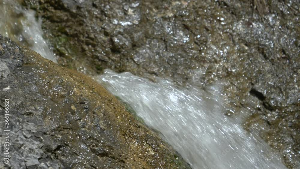 CLOSE UP, DOF: Glassy waterfall washes the dark brown and black stones on a sunny day. Detailed view of crystal clear stream water rushing down shiny rocks of a waterfall in Logar valley, Slovenia.
