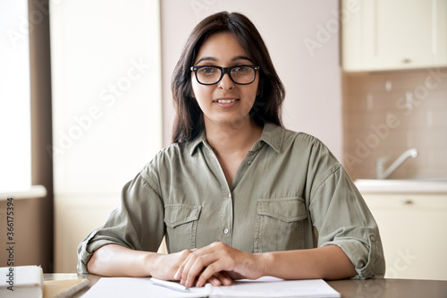 Smiling young indian female teacher wearing glasses sitting at table. Indian woman school tutor or university student studying working from home office giving online remote classes concept. Portrait