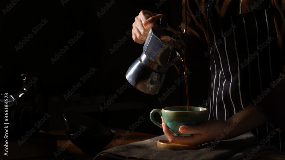 Female barista spills hot coffee from coffee pot to cup in dark background