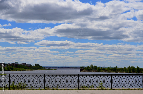 Fence on the embankment by the river under the blue sky with clouds