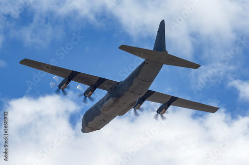 Large C-130 cargo plane used by the military flying against a partly cloudy sky.