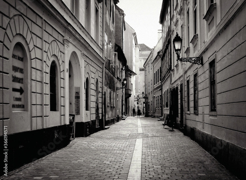 Narrow alley in old part of downtown in Gyor  Hungary with stucco exterior facades and arched windows in monochrome. cobblestone pavement. tourists walking in a distance. vintage wrought iron sconce