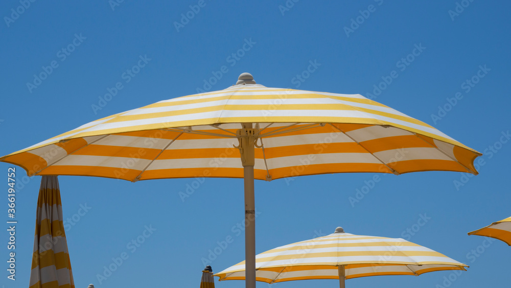 White and yellow striped beach umbrellas. Blue sky. Relaxing context. Summer holidays at the sea. General contest and location