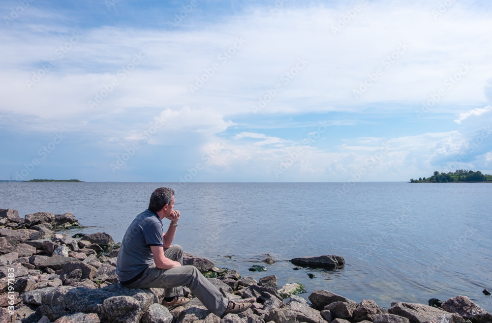 A lonely middle-aged man sits on a rocky beach on a summer day and looks thoughtfully into the distance. Loneliness, unity with nature concept. Horizontal orientation, selective focus.