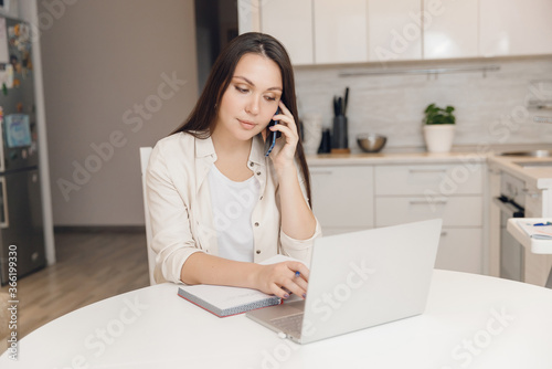 Young woman works in call center from office of her home kitchen. Calling customers through phone and filling out tables on laptop