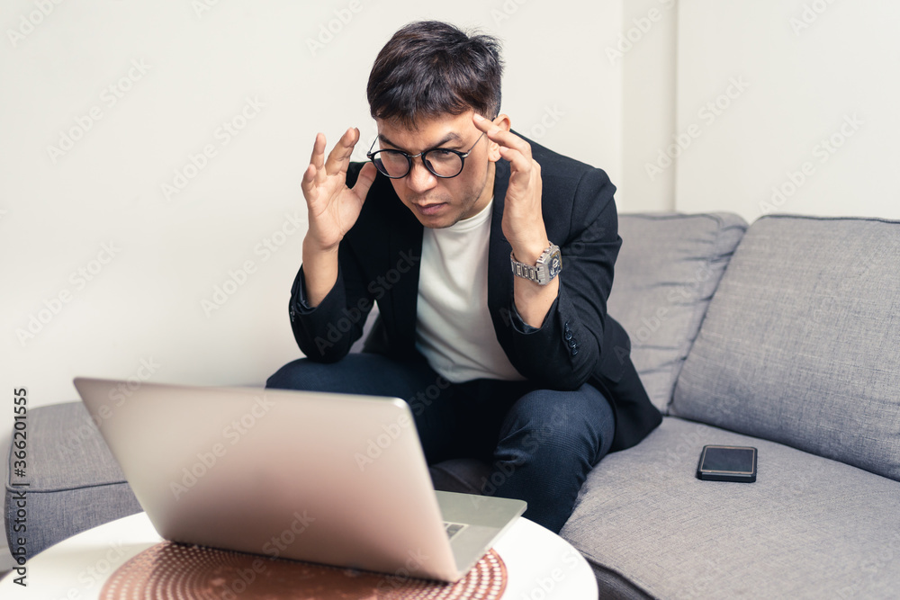 Young Asian business man sitting on sofa and working from home. Man feeling stress and suffering from headache. Tired young entrepreneur manager. Work from home concept Young Asian business man sitting on sofa and working from home. Man feeling stress and suffering from headache. Tired young entrepreneur manager. Work from home concept