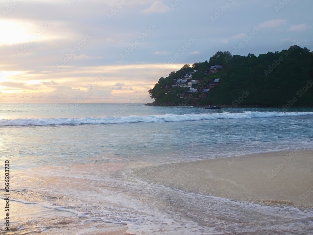 Tropical beach in Thailand. Sea, waves, small boat on the water on a distance. Buildings on the hill on the horizon. Residential buildings on a green slope. White sand, view of the bay, sunset time