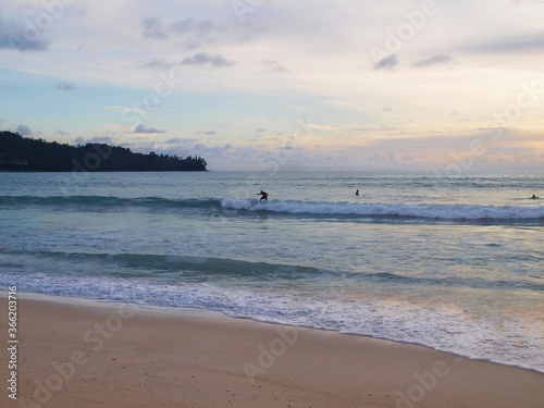 Surfer on the beach, water sport activity, sea waves. Surfing on the wave in twilight evening time. Popular resort, active rest, people. Sunset on the sea, beautiful sky with clouds.