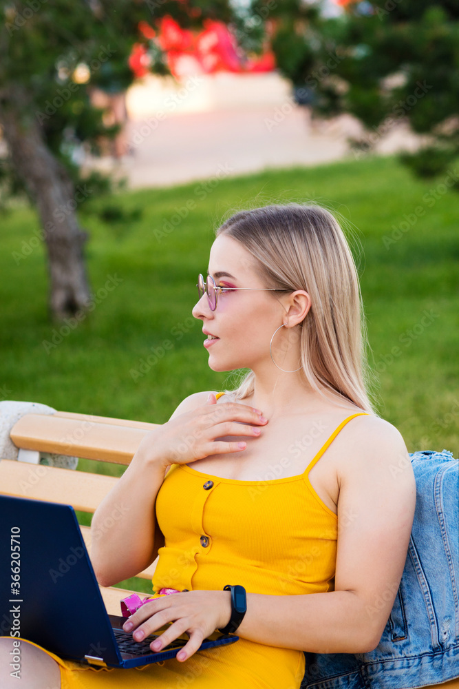 Obraz premium Young attractive girl in yellow dress works at laptop while sitting on bench in park. Concept of remote work from anywhere in the pestilence. Convenience. Freelance Worker.