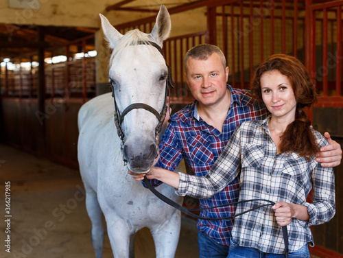 Mature smiling couple of farmers standing with white horse at stabling indoor