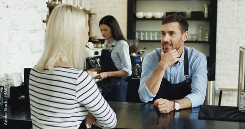 Handsome waiter talking with pretty blonde client at the bar, while waitress serving her coffee.