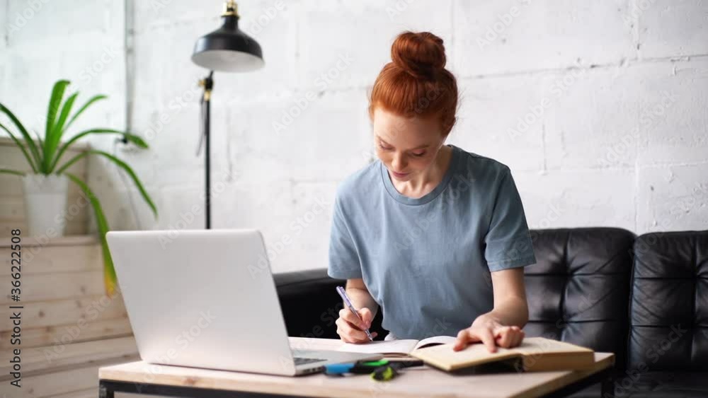 Beautiful redhead young woman student is noting into the workbook important information from the book in preparation for the lessons at table with laptop. Girl doing paperwork with book in home office Beautiful redhead young woman student is noting into the workbook important information from the book in preparation for the lessons at table with laptop. Girl doing paperwork with book in home office