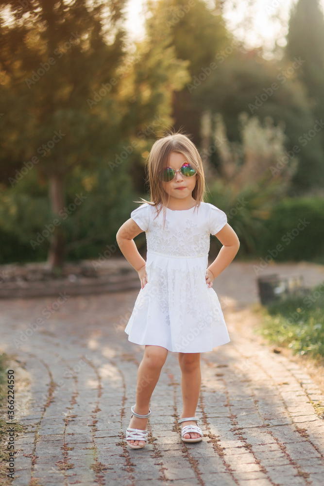 Cute little girl in white dress posing to photographer. Happy little kid in sunglasses