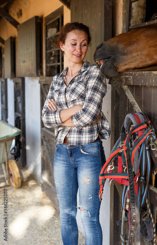 Mature positive female farmer standing near horse at stable outdoor