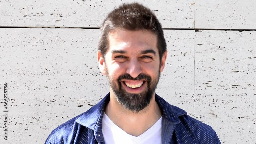 Close-up portrait of good-looking bearded guy in stylish clothing standing outside with straight face then smiling and looking at camera. Close-up portrait of good-looking bearded guy in stylish clothing standing outside with straight face then smiling and looking at camera.