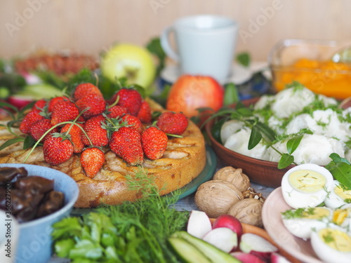Fragment of a table with vegetarian simple healthy food, crumpets, potatoes, eggs, herbs on a background of a cake with strawberries. Thanksgiving holiday table.