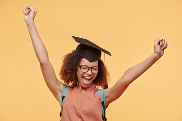 Happy excited black graduate student in eyeglasses and academic cap raising arms and screaming after completing her degree