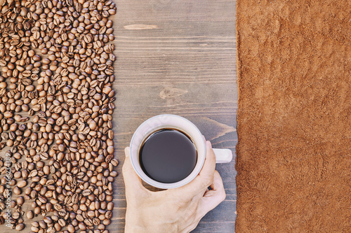 A top view of a person holding a cup of coffee with roasted and ground coffee beans on the table